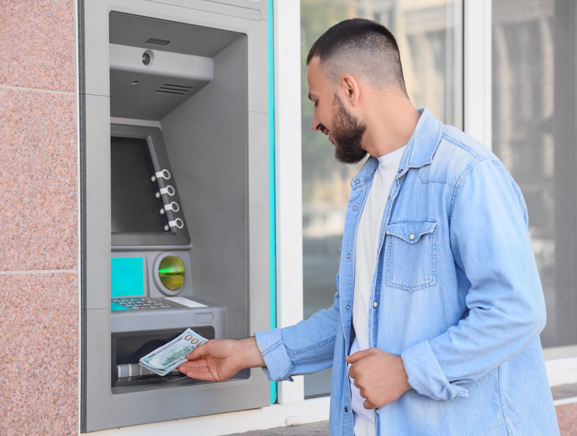 A smiling man in a denim shirt withdraws cash from an outdoor automated teller machine, or ATM.