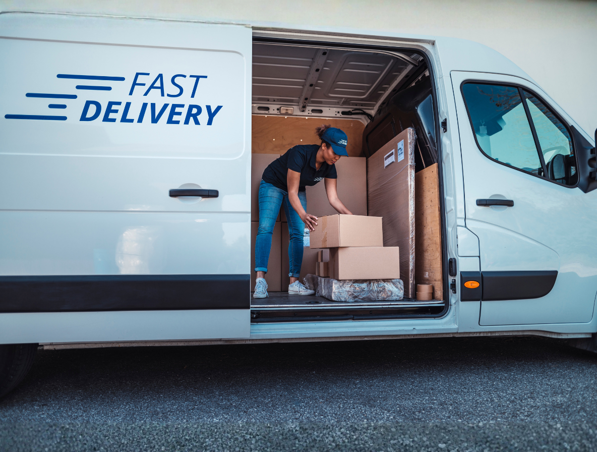 A delivery worker arranges cardboard boxes inside a white van labeled with a "FAST DELIVERY" logo.