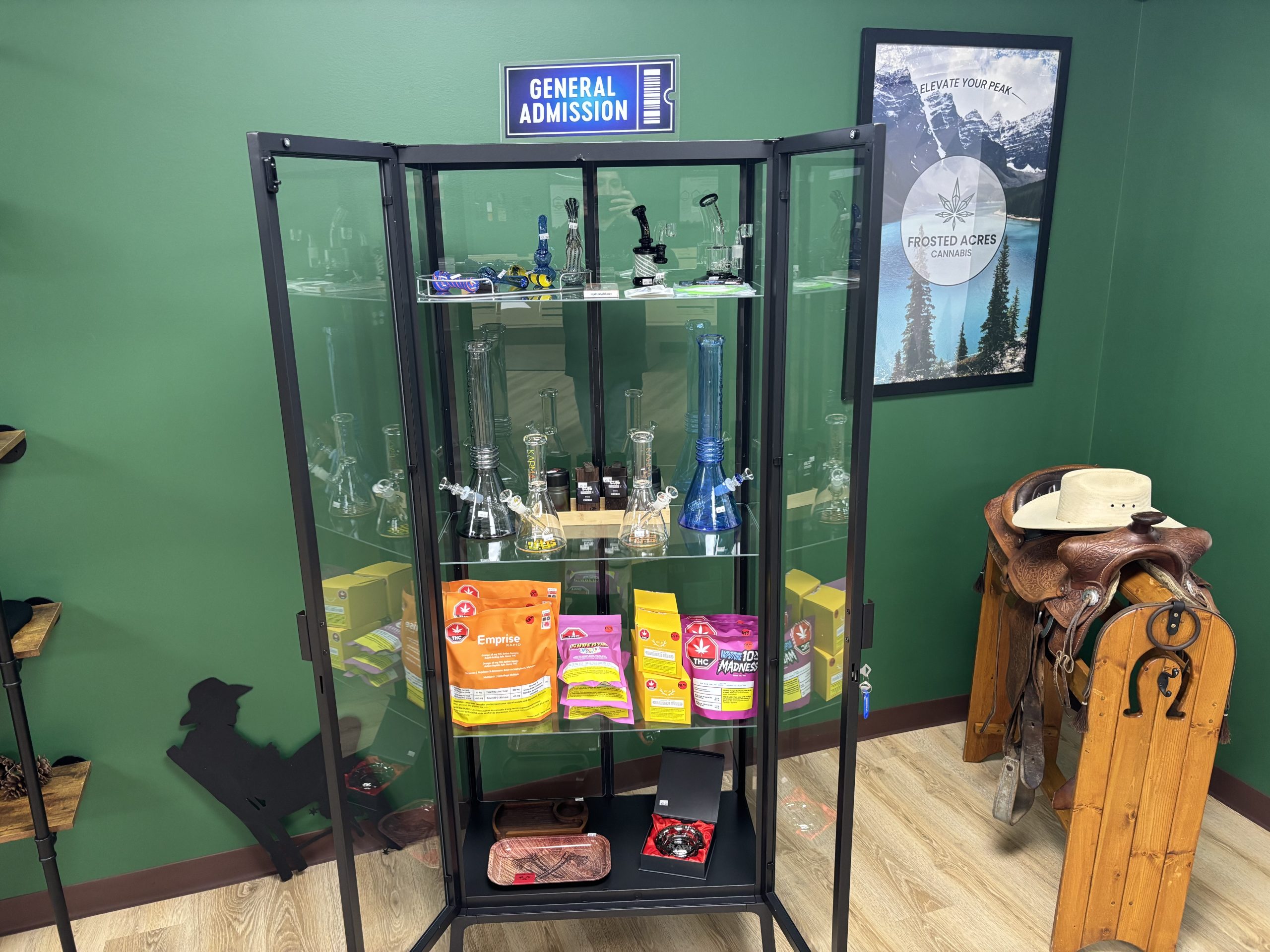 Glass display cabinet filled with cannabis products and bongs, next to a saddle with a cowboy hat.