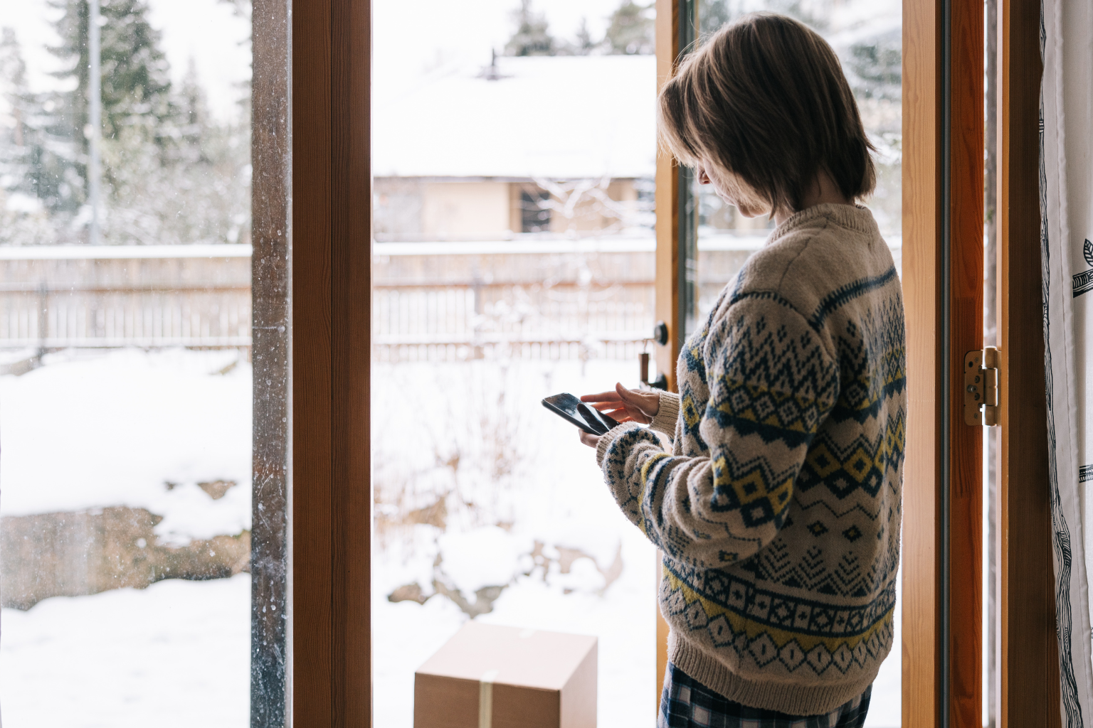 A woman in a patterned winter sweater checking their smartphone while standing by an open glass door, with a cardboard delivery box on the floor and a snowy yard outside.